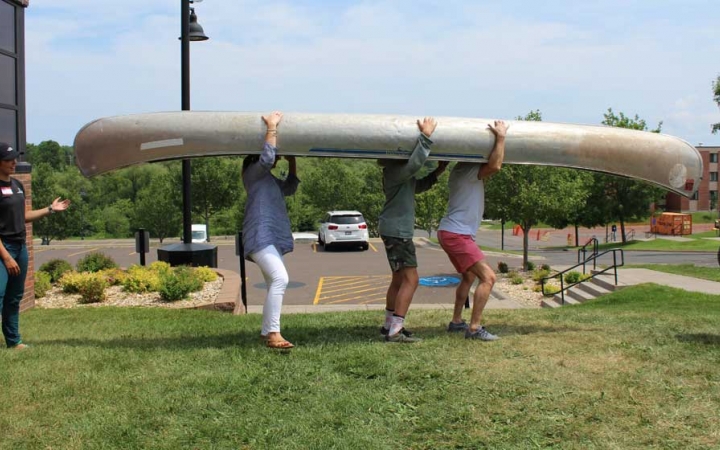 Three people carry an upside-down canoe on their shoulders at the family seminar of an outward bound intercept course